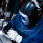 Person in blue sweater, white gloves and welding helmet performing a TIG weld on a Pedigo AGV cart