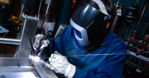 Person in blue sweater, white gloves and welding helmet performing a TIG weld on a Pedigo AGV cart
